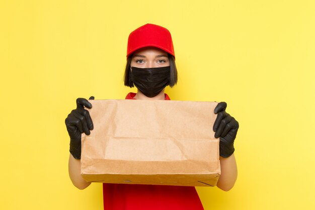 A front view young female courier in red uniform black gloves and red cap holding food package