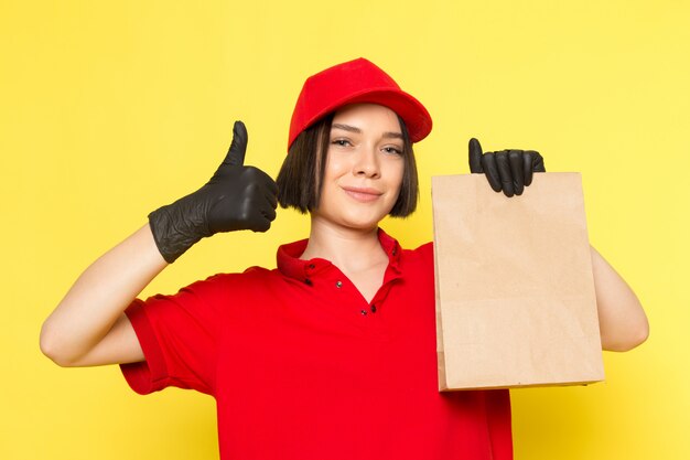 A front view young female courier in red uniform black gloves and red cap holding food package