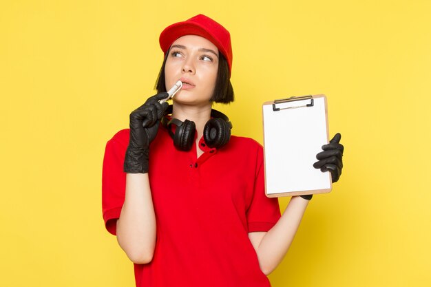 A front view young female courier in red uniform black gloves and red cap holding pen and notepad with thinking expression