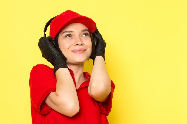 A front view young female courier in red uniform black gloves and red cap listening to music with delighted expression