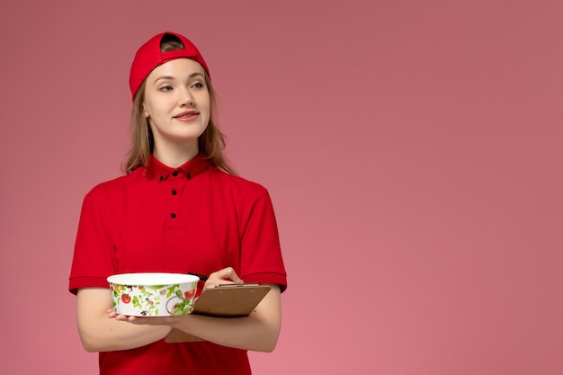 Front view young female courier in red uniform and cape holding delivery bowl and notepad on light pink wall