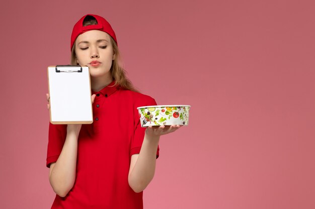 Front view young female courier in red uniform and cape holding delivery bowl and notepad thinking on pink wall