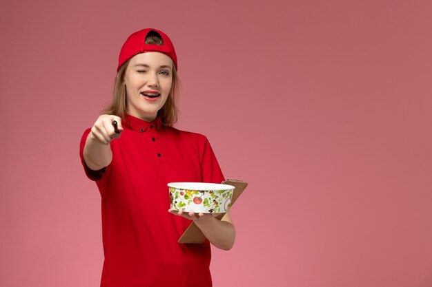 Front view young female courier in red uniform and cape holding delivery bowl and notepad with pen winking on pink wall