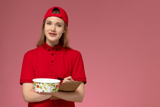 Front view young female courier in red uniform and cape holding delivery bowl and notepad writing notes on pink desk worker service uniform delivery