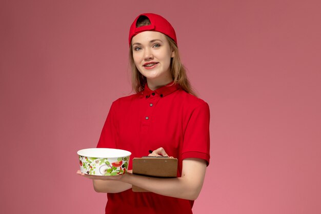 Front view young female courier in red uniform and cape holding delivery bowl and notepad writing notes on pink wall