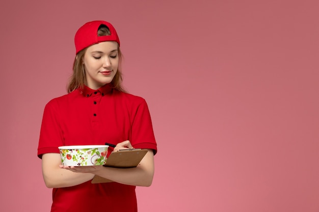 Front view young female courier in red uniform and cape holding delivery bowl and notepad writing notes on the pink wall