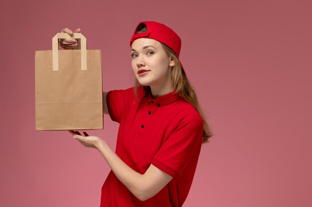Вид спереди молодая женщина-курьер в красной форме и накидке, держащая посылку с доставкой на розовой стене