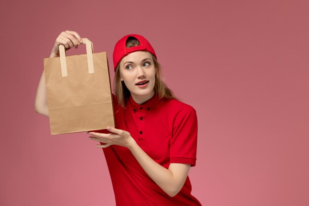 front view young female courier in red uniform and cape holding delivery food package on the pink wall
