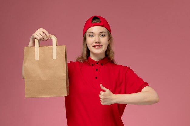 ピンクの壁に微笑んで配達食品パッケージを保持している赤い制服と岬の正面図若い女性の宅配便