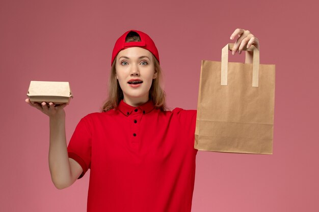 ピンクの壁に配達食品パッケージを保持している赤い制服と岬の正面図若い女性の宅配便