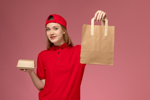 Вид спереди молодой женщины-курьера в красной форме и накидке, держащей пакеты с доставкой на розовой стене