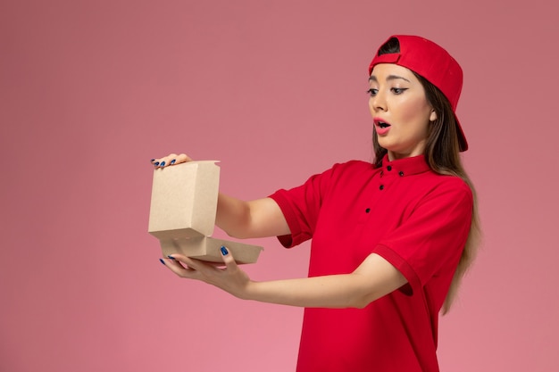Front view young female courier in red uniform and cape with little delivery food package on her hands on light pink wall