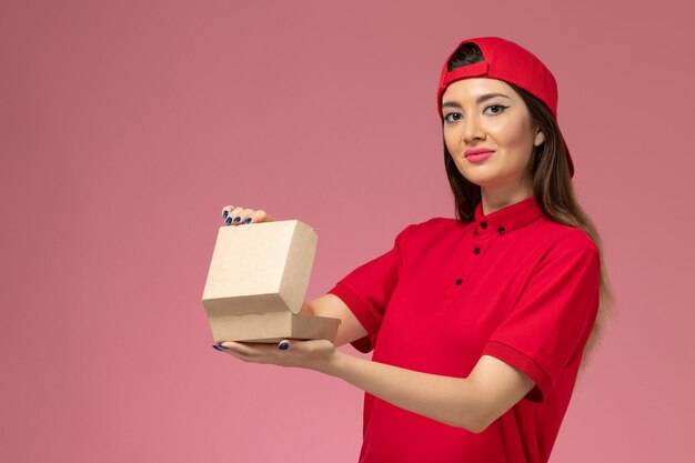 Front view young female courier in red uniform and cape with little delivery food package on her hands on light pink wall