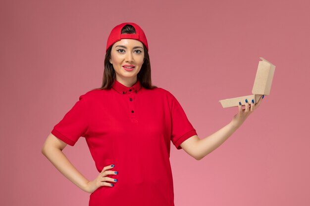 明るいピンクの壁に彼女の手に小さな配達食品パッケージと赤い制服とケープの正面図若い女性の宅配便
