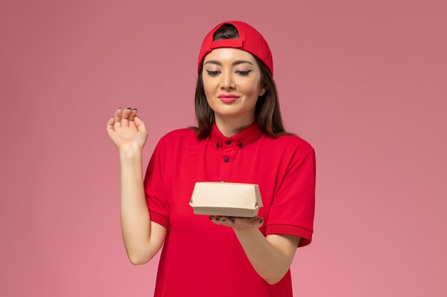 Front view young female courier in red uniform cape with little delivery food package on her hands on light pink wall
