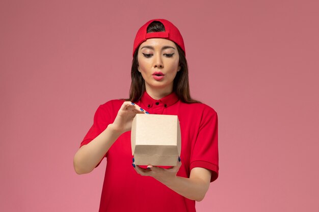 Front view young female courier in red uniform and cape with little delivery food package on her hands opening it on light pink wall