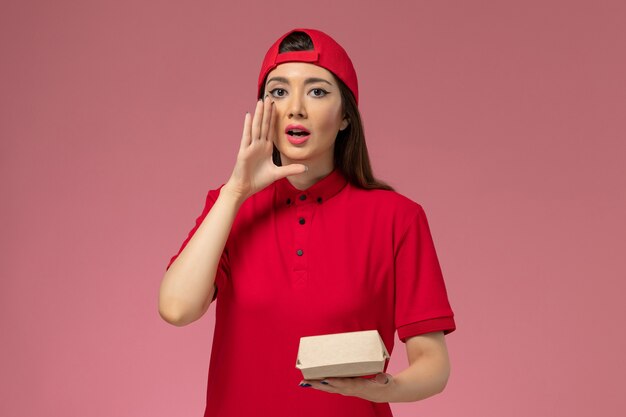 분홍색 책상 유니폼 배달 서비스에 그녀의 손에 작은 배달 음식 패키지와 빨간 유니폼과 케이프 전면보기 젊은 여성 택배