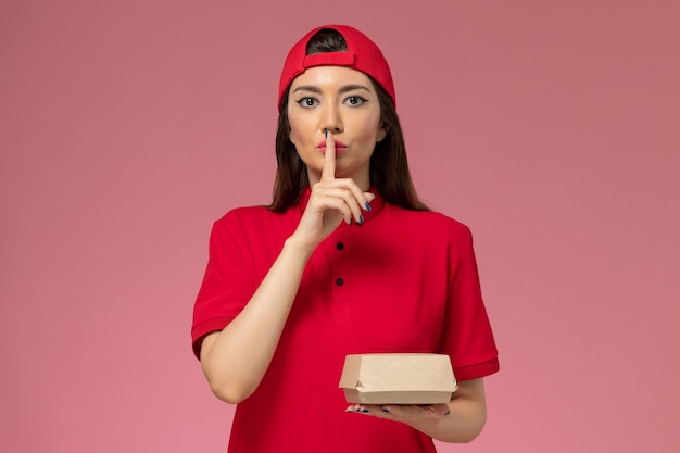 Foto gratuita corriere femminile giovane vista frontale in uniforme rossa e mantello con poco pacchetto di cibo di consegna sulle sue mani sul muro rosa