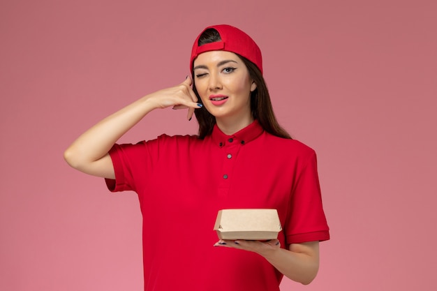 Front view young female courier in red uniform and cape with little delivery food package on her hands on pink wall