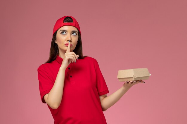 Front view young female courier in red uniform and cape with little delivery food package on her hands on the pink wall