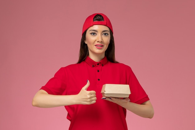 분홍색 벽에 그녀의 손에 작은 배달 음식 패키지와 빨간 유니폼과 케이프 전면보기 젊은 여성 택배