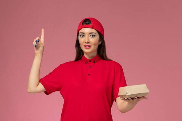 Front view young female courier in red uniform and cape with little delivery food package on her hands on the pink wall