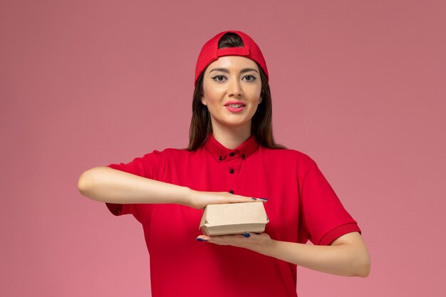 Front view young female courier in red uniform and cape with little delivery food package on her hands on the pink wall