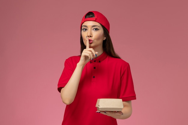 Молодая женщина-курьер в красной форме и накидке с маленьким пакетом еды на руках, показывая знак тишины на розовой стене, вид спереди
