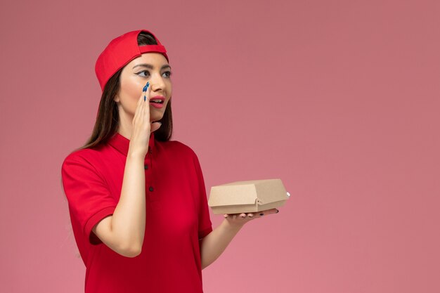Front view young female courier in red uniform and cape with little delivery food package on her hands and whispering on pink wall