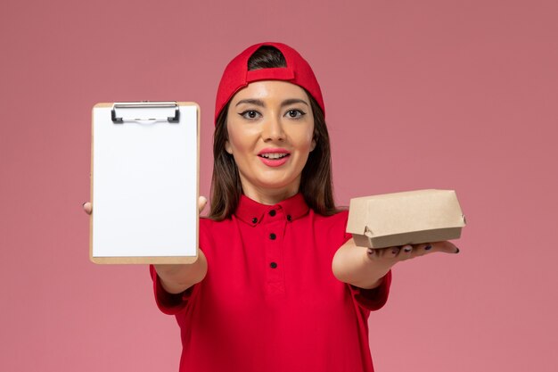 밝은 분홍색 벽에 그녀의 손에 작은 배달 음식 패키지와 메모장 빨간색 유니폼 케이프 전면보기 젊은 여성 택배