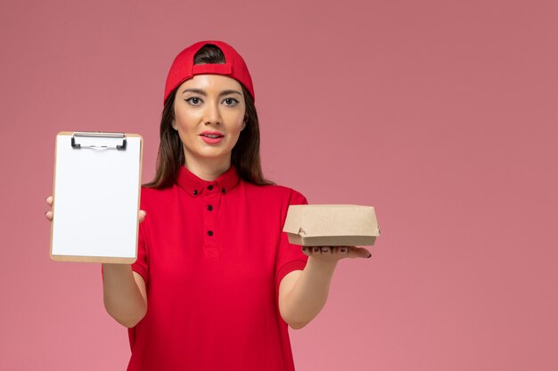 Front view young female courier in red uniform cape with little delivery food package and notepad on her hands on pink wall