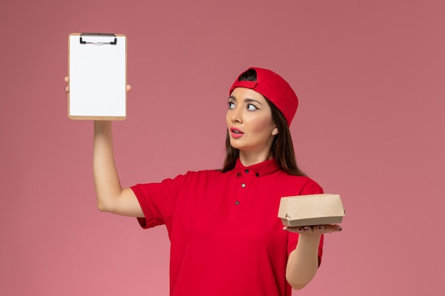 Giovane corriere femminile di vista frontale in mantello rosso uniforme con poco pacchetto di cibo di consegna e blocco note sulle sue mani sulla parete rosa