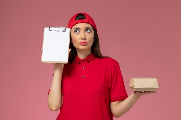 분홍색 벽에 그녀의 손에 작은 배달 음식 패키지와 메모장 빨간색 유니폼 케이프 전면보기 젊은 여성 택배