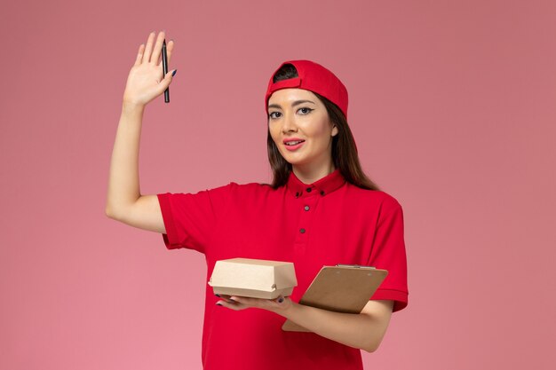 Corriere femmina giovane vista frontale in mantello rosso uniforme con piccolo pacchetto di cibo per la consegna e blocco note con la penna sulle mani sul muro rosa chiaro