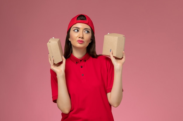 明るいピンクの壁に彼女の手に小さな配達食品パッケージと赤い制服とケープの正面図若い女性の宅配便