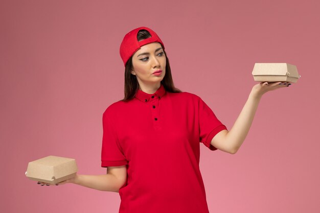 Вид спереди молодая женщина-курьер в красной форме и плаще с небольшими пакетами еды для доставки на руках на розовой стене