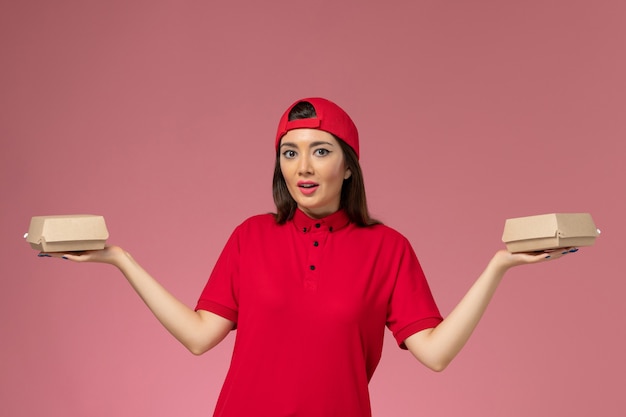 ピンクの壁に彼女の手に小さな配達食品パッケージと赤い制服とケープの正面図若い女性の宅配便