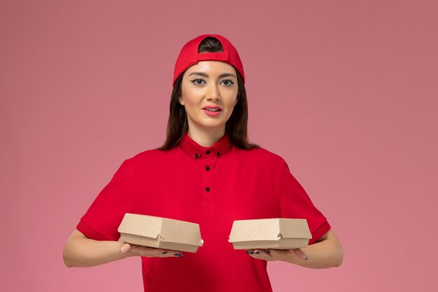 Front view young female courier in red uniform and cape with little delivery food packages on her hands on the pink wall