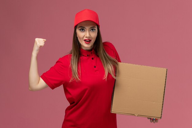 Front view young female courier in red uniform holding delivery food box and cheering on the pink background service delivery uniform company