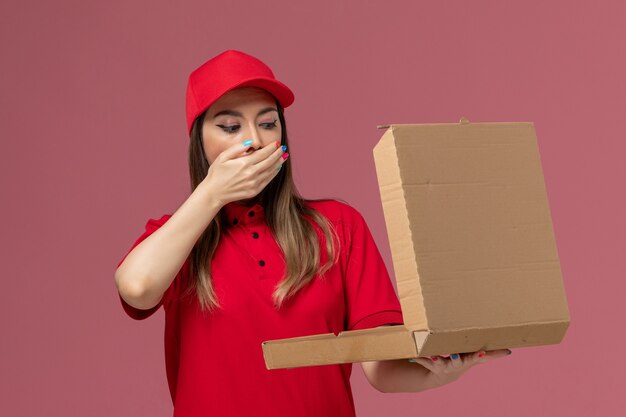 Front view young female courier in red uniform holding delivery food box and opening it on light-pink background service delivery job uniform company