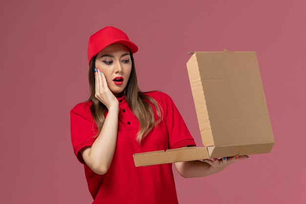 Front view young female courier in red uniform holding delivery food box opening it on the light-pink background service delivery uniform company