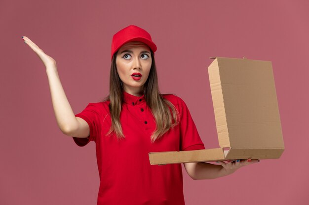 Giovane corriere femminile di vista frontale in uniforme rossa che tiene la scatola dell'alimento di consegna e aprendolo sulla società dell'uniforme di lavoro di consegna del servizio di scrivania rosa chiaro