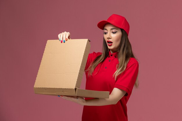 Front view young female courier in red uniform holding delivery food box and opening it on pink background service delivery job uniform company