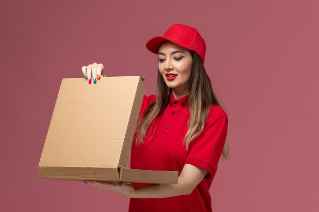 Front view young female courier in red uniform holding delivery food box opening it on pink background service delivery job uniform company
