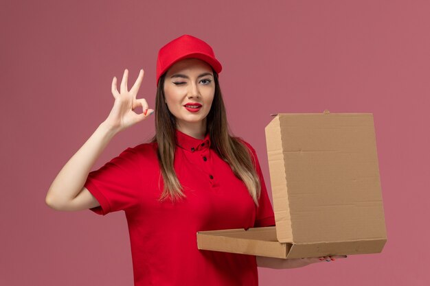 ピンクの背景にポーズをとって配達フードボックスを保持している赤い制服の正面図若い女性の宅配便配達仕事制服会社
