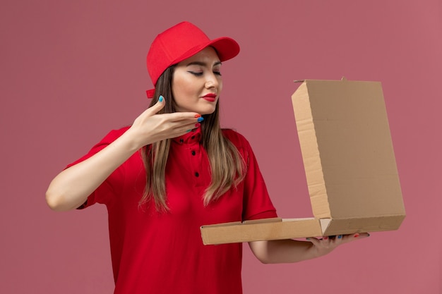 Front view young female courier in red uniform holding delivery food box smelling on light-pink background service delivery uniform worker company