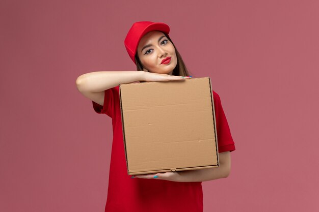 Вид спереди молодая женщина-курьер в красной форме, держащая коробку с едой для доставки, улыбаясь на розовом фоне.