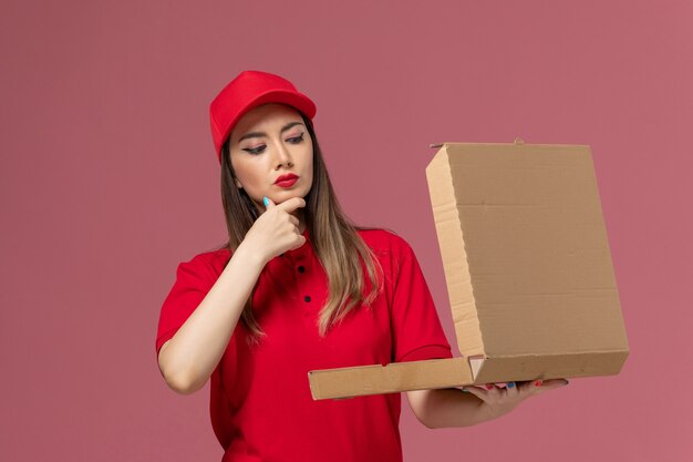 Front view young female courier in red uniform holding delivery food box thinking on light-pink background service delivery job uniform company