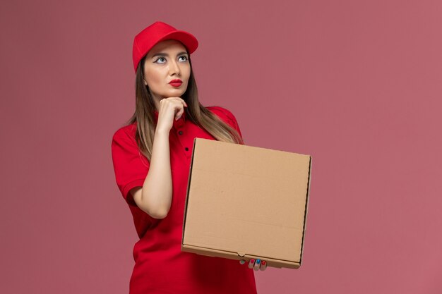 Вид спереди молодая женщина-курьер в красной форме, держащая коробку с едой для доставки, думая на розовом фоне, служба доставки униформы
