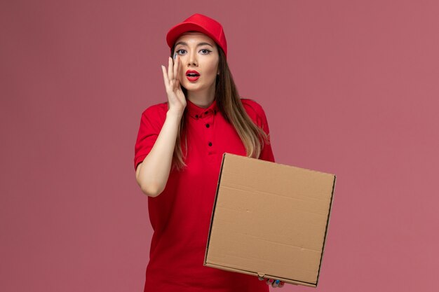 Front view young female courier in red uniform holding delivery food box whispering on the pink background service delivery uniform company